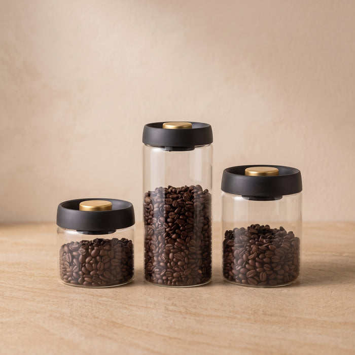 Three clear glass canisters filled with dark brown coffee beans, each with a black lid and a round gold-toned button, arranged on a light wood surface against a warm beige background. 500ml, 900ml and 1800ml